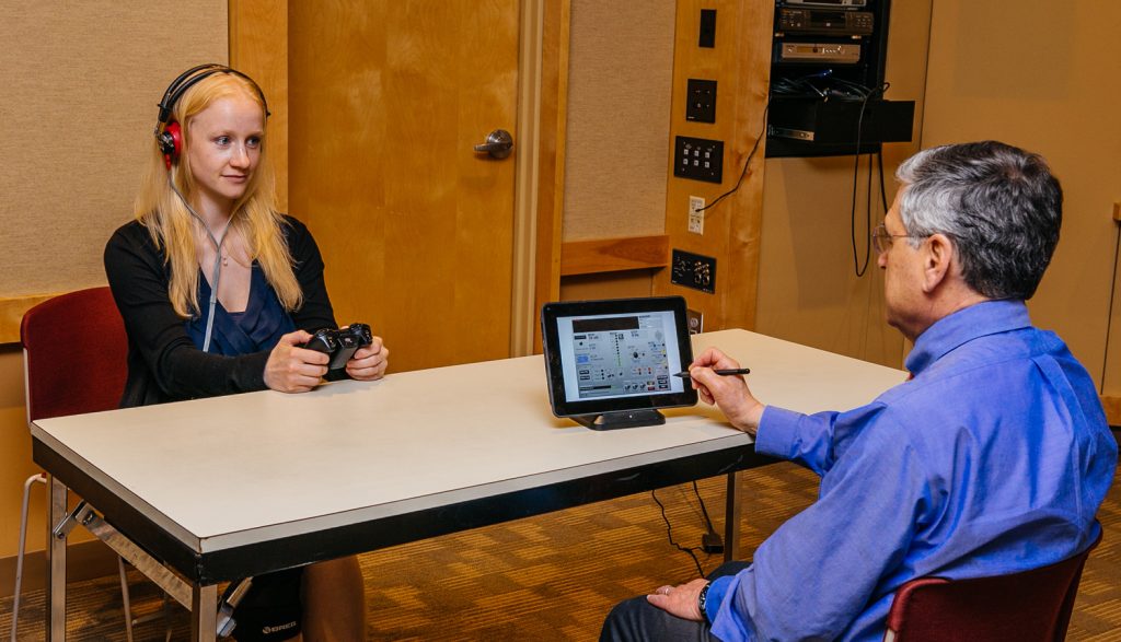 Tinnitus Diagnostic Tool Us Army, a women has headphones on and has an Xbox Controller in her hands, a researcher has a tablet in his hand opposite the table