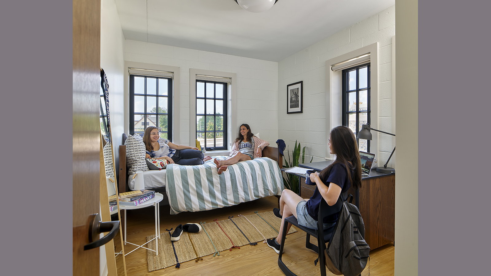 3 Villanova students enjoy their dormroom, two sit on the bed while one sits in a chair facing them,